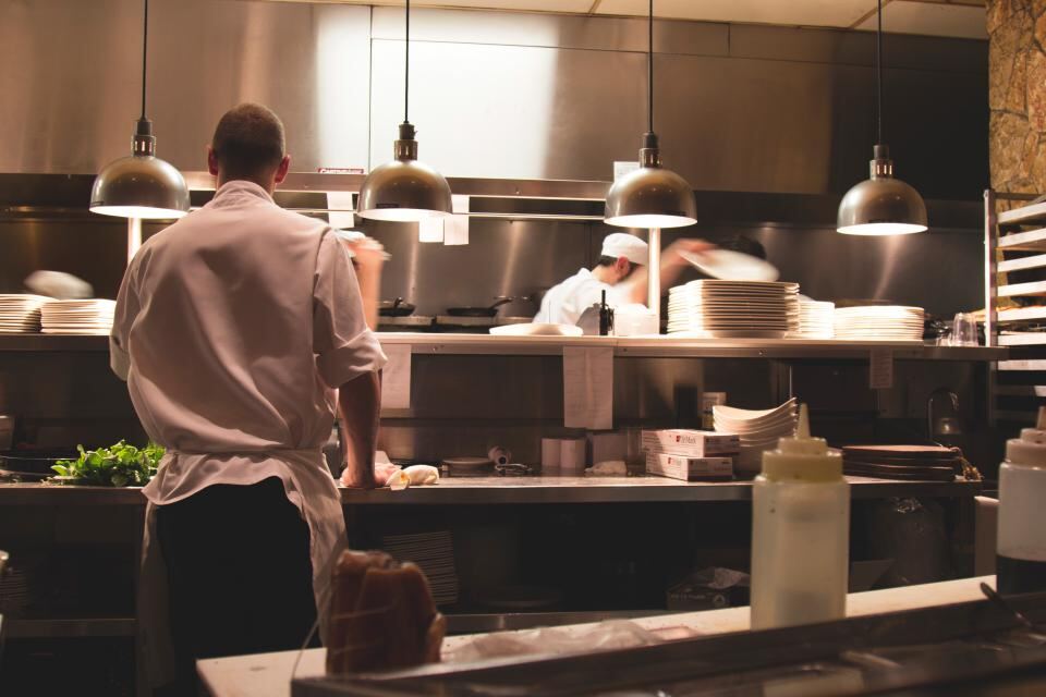 A chef in whites at the pass of a busy restaurant kitchen under warm pendant lights